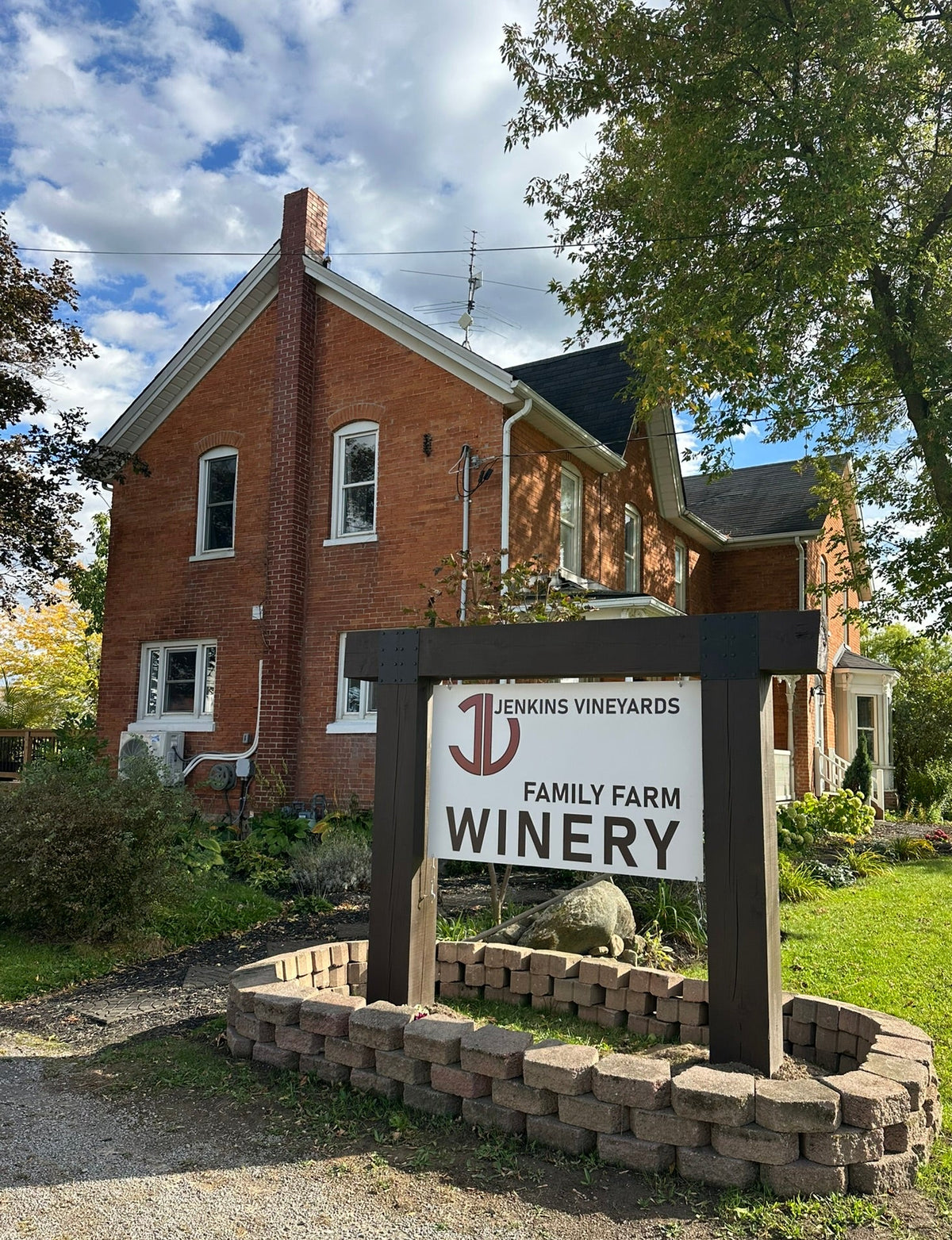 A brick house with a large wooden sign in front reading 'Jenkins Vineyards Family Farm Winery,' set in a landscaped yard with trees, grass, and a stone border.